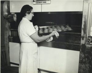 Image of a woman, dressed in white, removing baked goods from an oven.