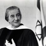 Black and white photograph of Golda Meir in a robe and seated in front of an Israeli flag.