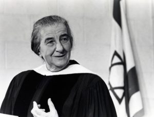 Black and white photograph of Golda Meir in a robe and seated in front of an Israeli flag.