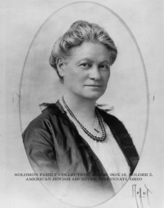 A black and white portrait photograph of Hannah G. Solomon, undated. Solomon is posed with her body turned sideways but her face looking forward, straight at the camera. She wears a formal jacket, a thick, beaded necklace, and her hair is in a bun. The image is shaped like an oval, as if retrieved from the inside of a locket.