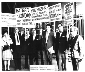 A black and white photograph of Rabbi Dr. Joachim Prinz picketing at the Jordanian Pavilion at the World’s Fair in New York, approx. April 1964. There are seven individuals visible in the image, six males and one female. Everyone is holding a picket sign. From left to right, the visible signs read: Number one, “Thou shalt not bear false witness against thy neighbor,” number two, “Hatred – Jordan, Help the refugees, don’t poison the…” number three, “King Hussein – Seek to promote international peace, not international hatred,” and number four, “Mid-east waters can give LIFE to all – Jordan – Make this possible.” Rabbi Dr. Prinz’s sign reads: “American Jewish Congress urges ‘Peace through understanding,’ Jordan incites war through bigotry.” The men are wearing suits with ties or bow ties, and Prinz adorns sunglasses. 