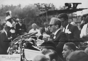 A black and white photograph of Rabbi Dr. Joachim Prinz giving a speech at the March on Washington, August 1963. He stands upon a raised platform in a crowd of men. He speaks into many microphones, at least five, from the podium on which he stands. Prinz wears sunglasses and a suit and tie, with an illegible button on the left collar. 
