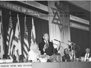 A black and white photograph of Rabbi Dr. Joachim Prinz on a panel with David Ben Gurion – the first Prime Minister of the State of Israel. Ben Gurion is sitting to the left, his face rested in his hand. Joachim Prinz is standing to Ben Gurion’s right, in front of three microphones, and appears mid-speaking. Behind them and the panel, on the back of the stage, are five flags alternating between the U.S. and Israeli flags, along with the words “[…] Conference on Israel” and “Jerusalem, June 1962” in English and the same phrases listed above them in Hebrew letters. In the center of the stage is a wall panel with the Star of David. 