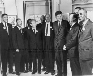 A black and white photograph of Rabbi Dr. Joachim Prinz [third from the left] with a group of men, including Martin Luther King, Jr [second from the left] and President Kennedy [third from the right]. The men are dressed in suits and dress clothes, with their arms around each other posing for the photograph. Each individual wears a matching round button, though the words or images on the button cannot be clearly made out. The individuals in this image led in the planning of the March on Washington. Washington, D.C., 1963. 